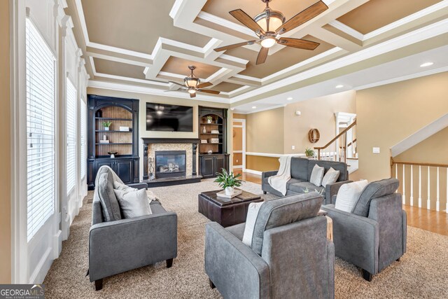 living area featuring ornamental molding, a glass covered fireplace, stairway, a ceiling fan, and coffered ceiling