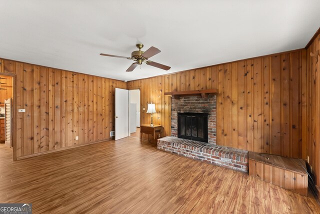 unfurnished living room with a ceiling fan, a brick fireplace, wood finished floors, and wooden walls