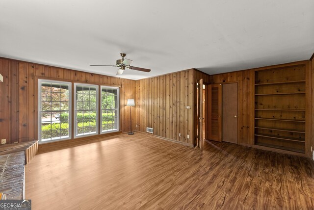 unfurnished living room featuring a ceiling fan, wood walls, built in shelves, and wood finished floors