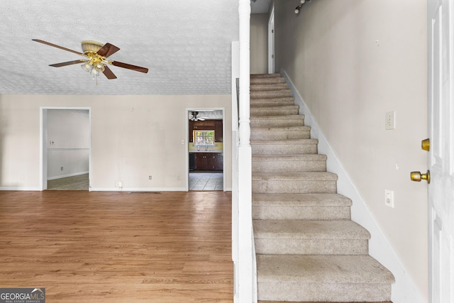 staircase with a textured ceiling, ceiling fan, baseboards, and wood finished floors