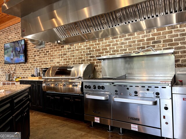 kitchen with wall chimney range hood, light stone counters, dark cabinets, finished concrete floors, and tasteful backsplash
