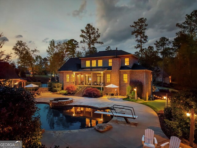 back of house at dusk featuring a patio, a pool with connected hot tub, and brick siding