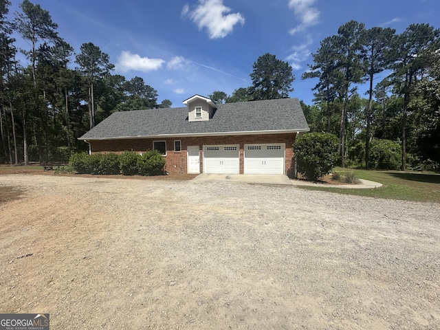 exterior space featuring a shingled roof, dirt driveway, an attached garage, and brick siding