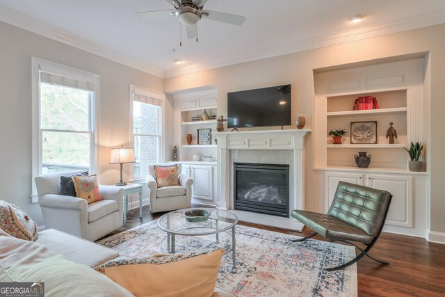 living room featuring crown molding, dark wood-style flooring, a glass covered fireplace, built in features, and a ceiling fan
