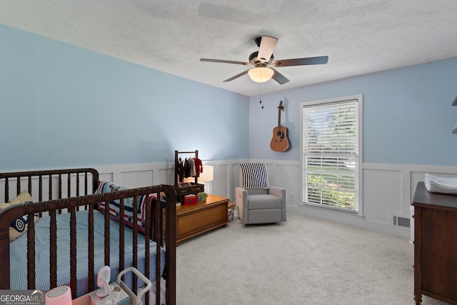 carpeted bedroom featuring a decorative wall, a textured ceiling, visible vents, ceiling fan, and wainscoting