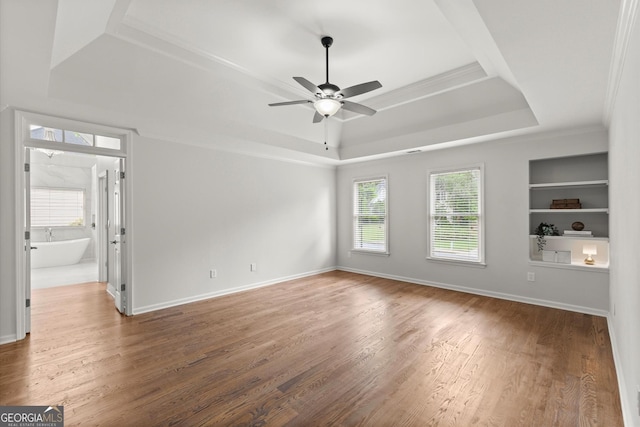 empty room featuring ceiling fan, built in features, wood finished floors, a tray ceiling, and ornamental molding