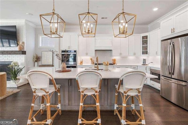kitchen with ornamental molding, glass insert cabinets, a center island with sink, light countertops, and appliances with stainless steel finishes