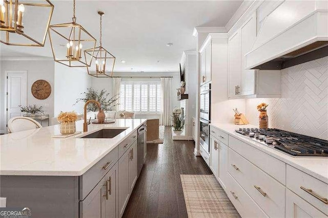 kitchen featuring a kitchen island with sink, custom range hood, a sink, appliances with stainless steel finishes, and a notable chandelier