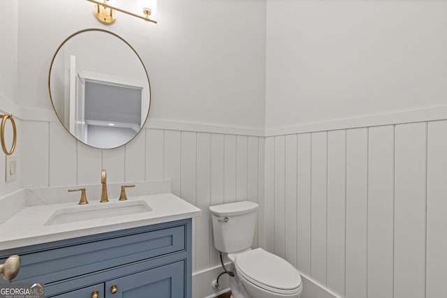 half bathroom with a decorative wall, toilet, a wainscoted wall, and vanity