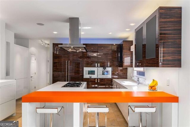 kitchen featuring island exhaust hood, black electric cooktop, stainless steel gas cooktop, a sink, and a breakfast bar