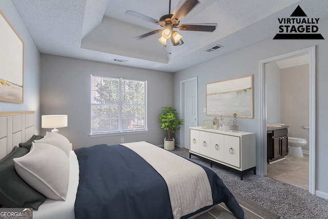bedroom featuring light carpet, ensuite bath, a raised ceiling, a ceiling fan, and a textured ceiling
