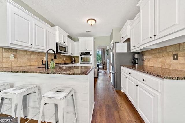 kitchen featuring appliances with stainless steel finishes, a peninsula, dark stone counters, a sink, and white cabinetry