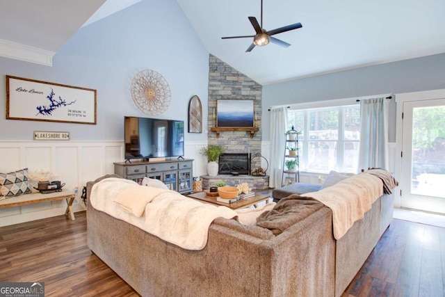 living area featuring a stone fireplace, high vaulted ceiling, dark wood-type flooring, a wainscoted wall, and a ceiling fan