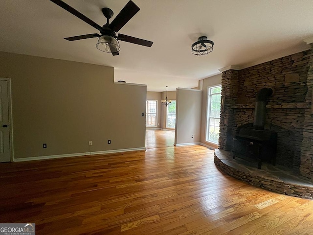 unfurnished living room featuring baseboards, a wood stove, ceiling fan with notable chandelier, and wood finished floors
