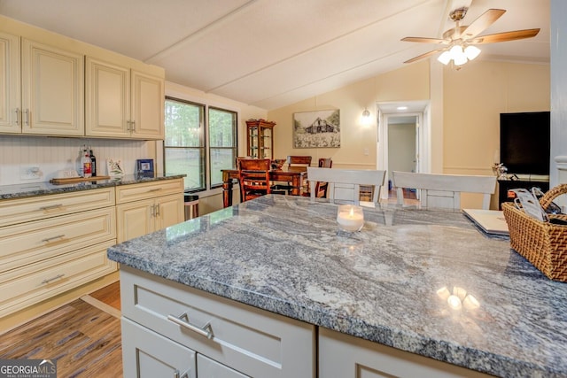 kitchen with a ceiling fan, cream cabinets, vaulted ceiling, wood finished floors, and stone countertops