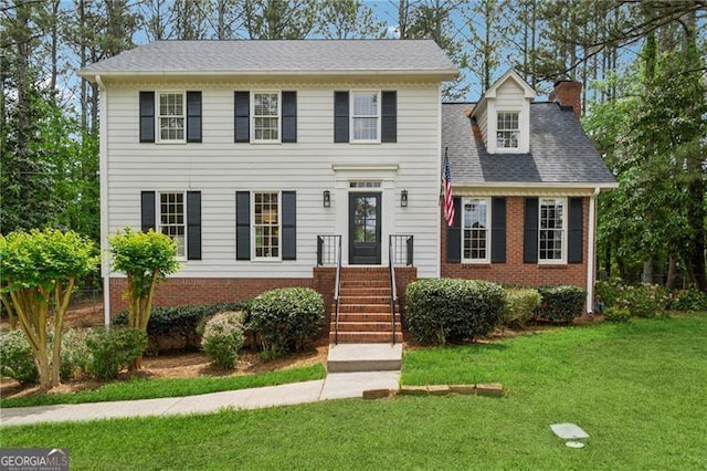 colonial home featuring a shingled roof, a chimney, a front yard, and brick siding