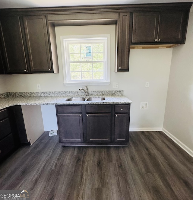 kitchen with dark wood-style flooring, dark brown cabinetry, light stone countertops, a sink, and baseboards