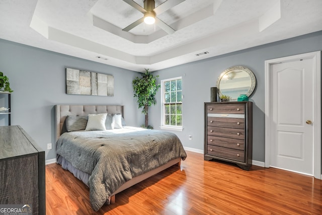 bedroom featuring a raised ceiling, a ceiling fan, baseboards, wood finished floors, and visible vents