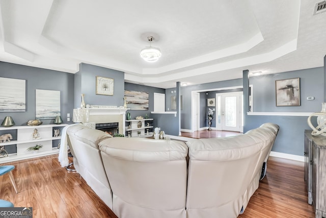 living area featuring visible vents, baseboards, wood finished floors, a tray ceiling, and a lit fireplace