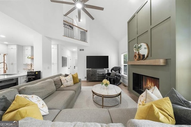living area with ceiling fan, visible vents, wood finished floors, high vaulted ceiling, and a warm lit fireplace