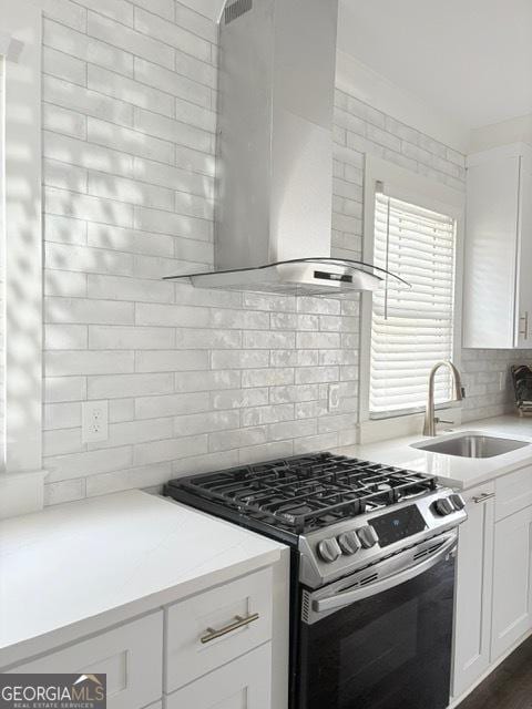 kitchen featuring stainless steel range with gas stovetop, wall chimney range hood, a sink, backsplash, and white cabinetry