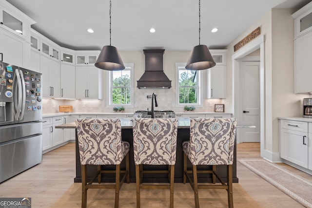 kitchen with decorative backsplash, a center island with sink, stainless steel fridge with ice dispenser, custom range hood, and light wood-style flooring