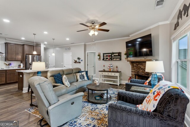 living area with ceiling fan, dark wood-style flooring, a stone fireplace, ornamental molding, and baseboards