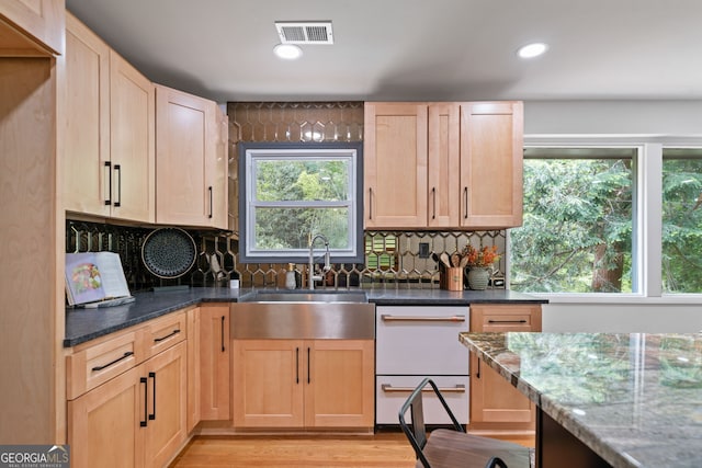 kitchen featuring light brown cabinetry, a sink, dark stone countertops, white dishwasher, and backsplash