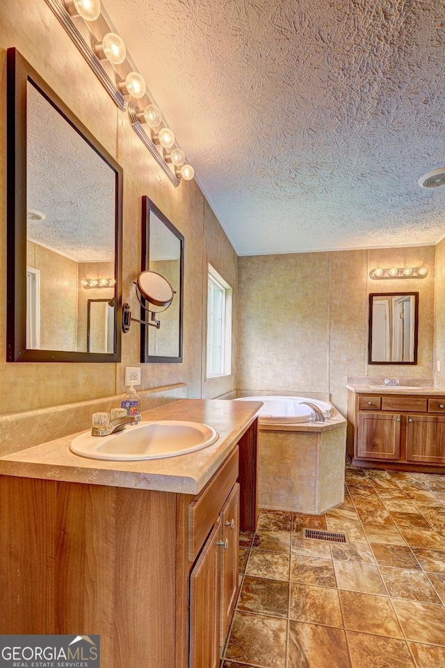 full bath featuring a textured ceiling, two vanities, a sink, and a garden tub