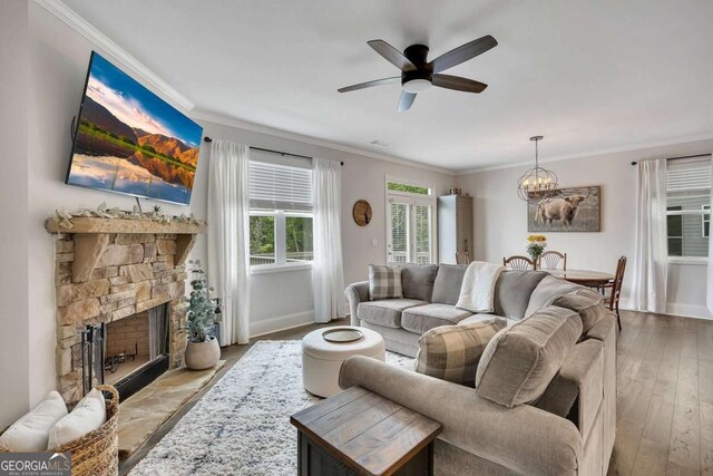 living area with a stone fireplace, ceiling fan with notable chandelier, baseboards, ornamental molding, and hardwood / wood-style flooring