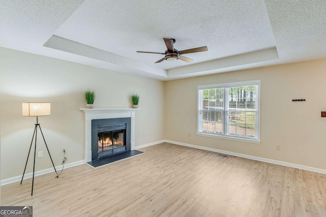 unfurnished living room with a ceiling fan, a textured ceiling, a fireplace with flush hearth, light wood-style floors, and a tray ceiling