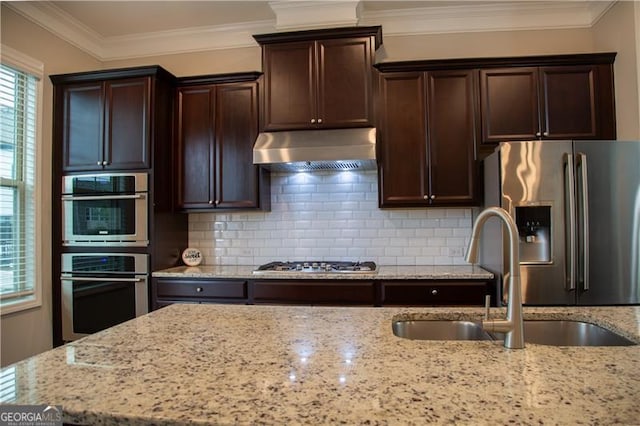 kitchen featuring under cabinet range hood, appliances with stainless steel finishes, a sink, light stone counters, and decorative backsplash