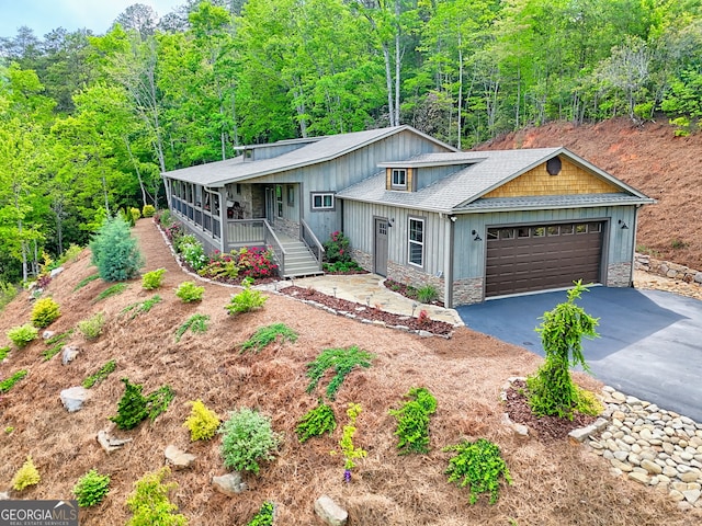 view of front facade with a porch, stone siding, driveway, an attached garage, and a wooded view