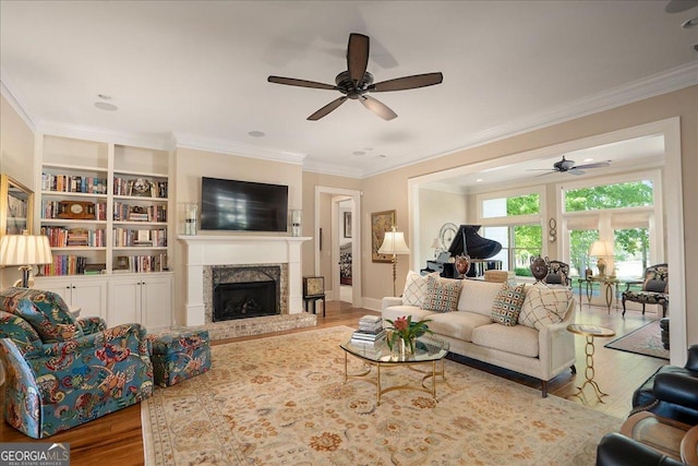 living area with a fireplace with raised hearth, ceiling fan, wood finished floors, and ornamental molding