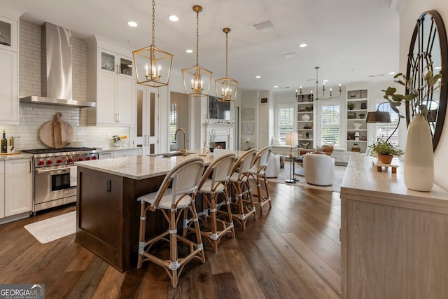 kitchen featuring a sink, wall chimney range hood, designer stove, a center island with sink, and an inviting chandelier