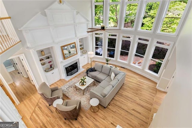 living room featuring ceiling fan, built in features, light wood-style flooring, a fireplace with flush hearth, and a towering ceiling