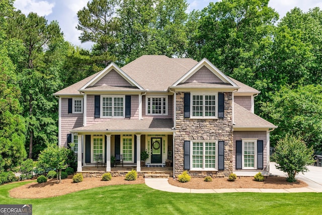 colonial house featuring covered porch, roof with shingles, stone siding, and a front lawn
