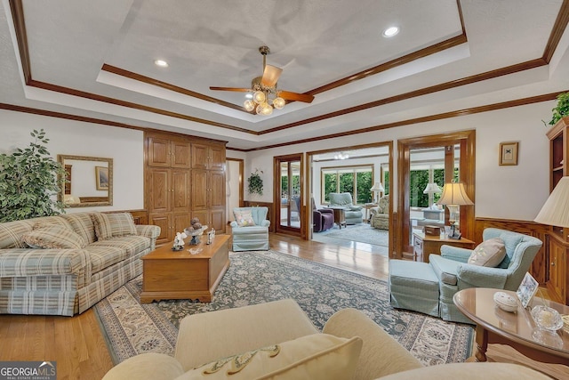 living room with wainscoting, light wood-style flooring, a ceiling fan, a tray ceiling, and ornamental molding