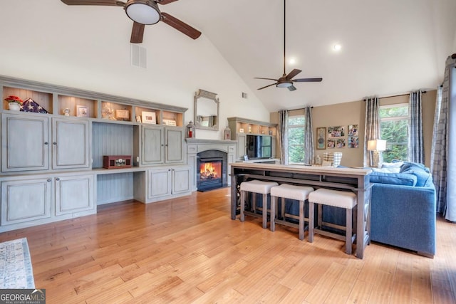 living room featuring a ceiling fan, high vaulted ceiling, a glass covered fireplace, and light wood finished floors