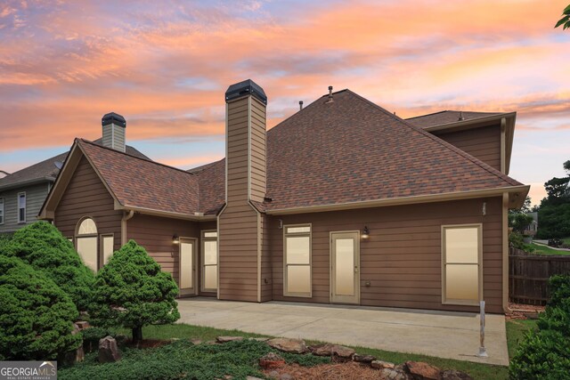 back of property at dusk with a shingled roof, a patio, a chimney, and fence