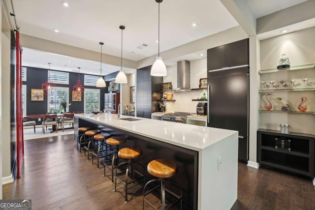 kitchen featuring wall chimney range hood, a sink, a breakfast bar area, light countertops, and dark wood-style floors