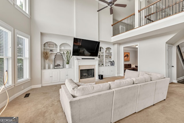 carpeted living area with a towering ceiling, built in features, a ceiling fan, baseboards, and a fireplace