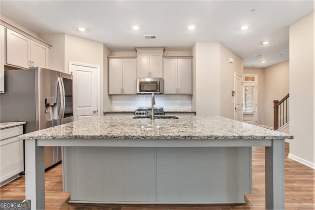 kitchen with light stone counters, a kitchen island with sink, light wood-style flooring, stainless steel appliances, and backsplash