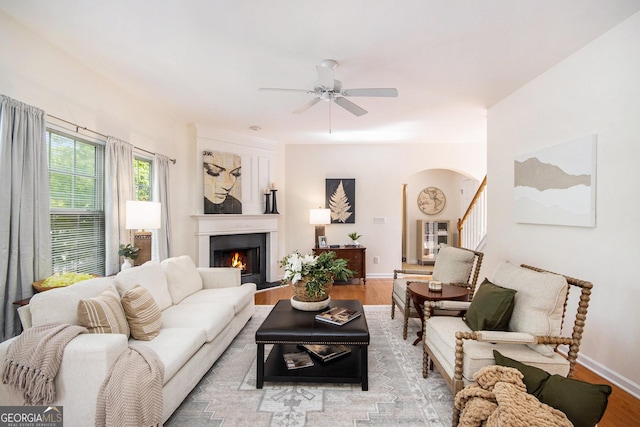 living room featuring arched walkways, a ceiling fan, wood finished floors, and a lit fireplace