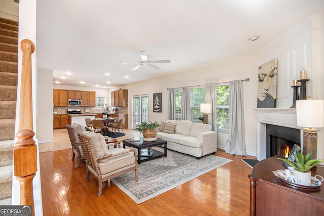 living room with ceiling fan, a warm lit fireplace, stairs, recessed lighting, and light wood-type flooring