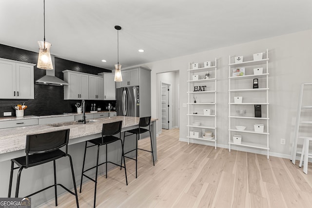 kitchen with a sink, light wood finished floors, a breakfast bar area, wall chimney exhaust hood, and freestanding refrigerator