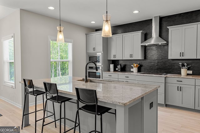 kitchen with a center island with sink, wall chimney range hood, tasteful backsplash, a sink, and appliances with stainless steel finishes