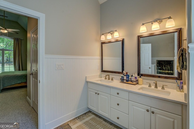 ensuite bathroom with a ceiling fan, a wainscoted wall, double vanity, lofted ceiling, and baseboards