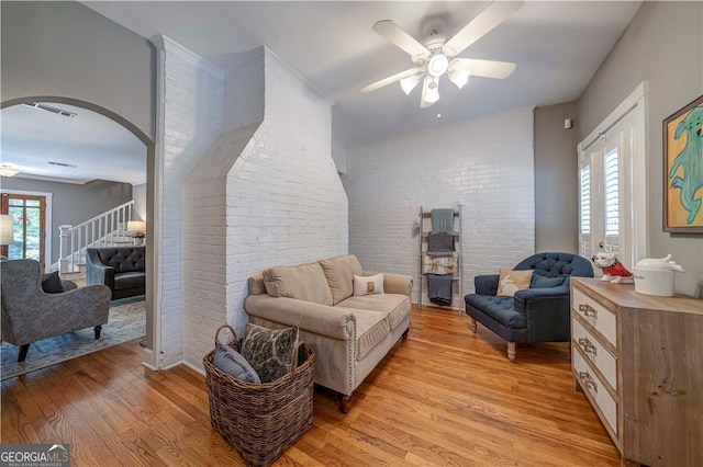 living room featuring brick wall, light wood-type flooring, ceiling fan, arched walkways, and stairs