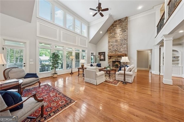 living room featuring a ceiling fan, light wood-style flooring, decorative columns, a fireplace, and a high ceiling